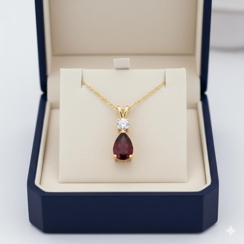 Puede incluir: Un collar dorado con un colgante de piedra preciosa roja oscura en forma de pera y una peque&ntilde;a piedra preciosa clara y redonda. El collar se exhibe en una caja de joyer&iacute;a azul con interior crema.