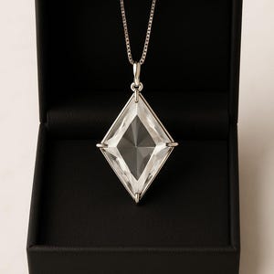 May include: A clear, diamond-shaped crystal pendant on a silver chain, displayed in a black box. The pendant is framed in silver. The necklace is a striking piece of jewellery, suitable for special occasions.