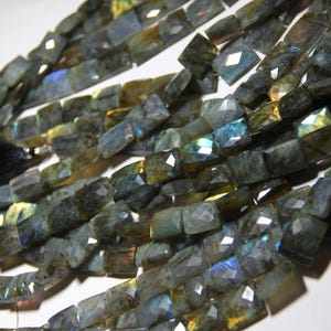 Pode incluir: Um fio de contas de pedra preciosa de labradorita cinza escuro e azul. As contas são facetadas e têm forma retangular. As contas são enfiadas em um cordão preto.