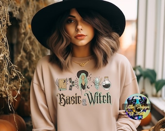 May include: A woman wearing a beige sweatshirt with the text "Basic Witch" and a graphic of a witch with a book, a crystal, a potion bottle, and a candle. The woman is wearing a black hat and has long brown hair.