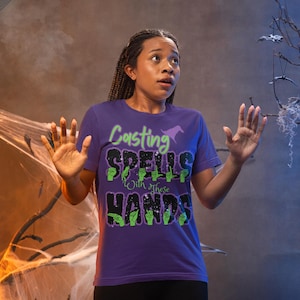 May include: A purple t-shirt with the text "Casting Spells With These Hands" in green and black. The shirt features a witch hat graphic. The person is wearing black leggings. The background includes a Halloween theme with cobwebs and a carved pumpkin.