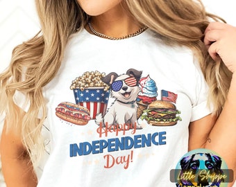 May include: White t-shirt with a cartoon dog wearing sunglasses and a red, white, and blue bandana. The dog is surrounded by American-themed food like hot dogs, hamburgers, ice cream, and popcorn. The text "Happy Independence Day!" is printed on the shirt.