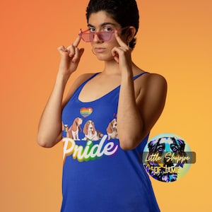 May include: A blue tank top featuring a rainbow heart and the word "Pride" in rainbow lettering. The tank top has a graphic of four basset hounds wearing sunglasses. The tank top is being worn by a person with short dark hair and pink glasses. The image also includes a round sticker with a dog graphic and the text "Little Shoppe of Jamie".