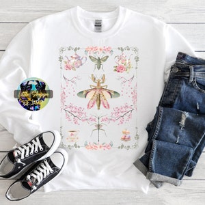 Könnte beinhalten: Weißer Pullover mit einem grafischen Druck von Insekten und Blumen in einem rosa und grünen Farbschema. Die Grafik ist mit einem floralen Rahmen eingefasst. Der Text "Little Shoppe of Jamie" ist in der linken unteren Ecke des Bildes sichtbar.