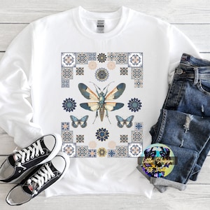 Puede incluir: Sudadera blanca con un patrón geométrico azul y dorado que presenta un insecto grande con detalles de mariposa. El texto "Little Shoppe of Jamie" es visible en la esquina inferior derecha.