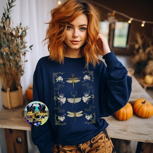 Könnte beinhalten: Ein marineblaues Sweatshirt mit einem grafischen Druck von drei stilisierten Insekten und floralen Akzenten. Die Insekten sind ein Stabheuschrecke, eine Libelle und eine Wasserjungfer. Der Text "Little Shoppe of Jamie" ist in einem Kreis unten links im Bild sichtbar.