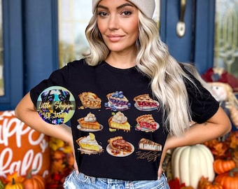 May include: A black t-shirt with a graphic of nine different pie slices. The slices include apple, blueberry, cherry, lemon meringue, strawberry, pecan, and cheesecake. The text "Little Shoppe of Jamie" is visible in the lower left corner of the image.