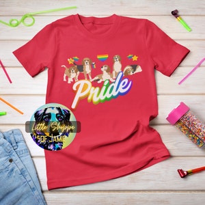 Puede incluir: Camiseta roja con un gráfico de arcoíris que dice "Pride" y un grupo de perros de dibujos animados. Los perros llevan collares de arcoíris y hay estrellas y corazones a su alrededor.