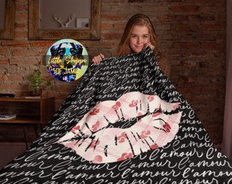 Può includere: Una coperta nera con un grande disegno di labbra a quadri rosa e bianche. La coperta ha le parole "l'amour" scritte in corsivo bianco. La coperta è tenuta da una persona.