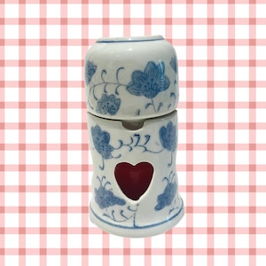 Puede incluir: Un quemador de aceite de cerámica blanca con un diseño floral azul. El quemador tiene una abertura en forma de corazón en el centro, que revela una fusión de cera roja.