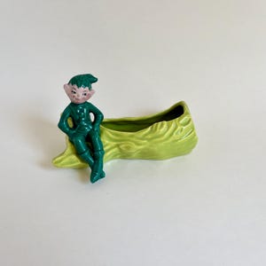 May include: A vintage ceramic planter featuring a seated green elf figure. The elf is perched on a light green, log-shaped planter. The planter has a hollowed-out interior for holding small plants or decorative items. The elf has a painted face.