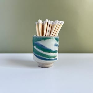 May include: A small, ceramic match holder with a blue, green, and white swirl design. The holder is filled with wooden matches with white tips. The holder sits on a white surface against a muted green background.
