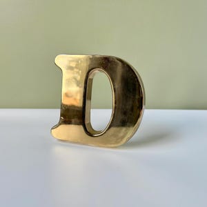 May include: A three-dimensional letter D with a polished gold-tone metallic finish. The letter features a smooth, reflective surface and a bold, block-style font design, standing upright with a hollow center opening.