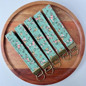 May include: Four fabric wristlet keychains featuring a sage green background with a repeating floral pattern in shades of pink, orange, and white. Each strap is finished with a rectangular antique brass metal clamp and a circular split key ring.