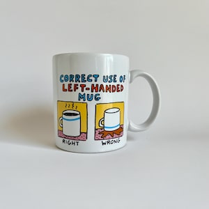 May include: White ceramic mug with a curved handle. The mug features the text "CORRECT USE OF LEFT-HANDED MUG" in blue and red, along with illustrations showing the right and wrong way to hold the mug.