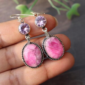 Puede incluir: Pendientes ovalados de color rosa con un pequeño strass rosa en la parte superior. Los pendientes son de plata y tienen un diseño delicado.