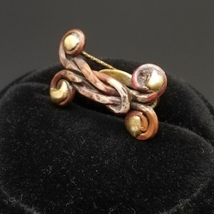 Puede incluir: Un anillo artesanal con un diseño único. El anillo presenta metales de color cobre, bronce y oro con un patrón abstracto en espiral. El anillo tiene un aspecto texturizado y está sobre un fondo oscuro.