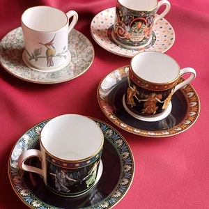 May include: Four decorative teacups and saucers with various designs. One cup features a hummingbird, another has a floral pattern, and two have classical figures. The saucers have coordinating patterns and gold trim, set against a red background.