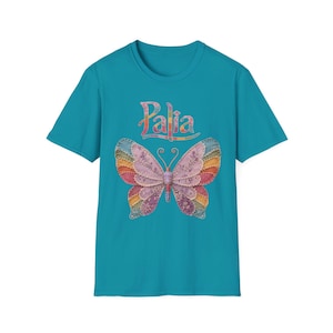 May include: A teal short-sleeved t-shirt featuring the word Palia in a multicoloured, patterned font above a large, symmetrical butterfly graphic. The butterfly wings are composed of various floral and geometric fabric-like textures in shades of pink, purple, orange, and blue.