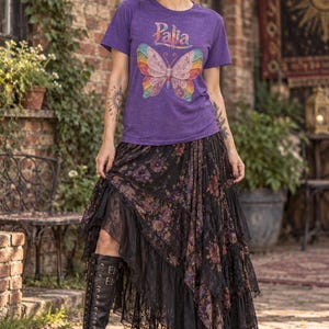 May include: A purple short-sleeved t-shirt featuring the word Palia in colorful lettering above a large, multi-colored butterfly graphic. The shirt is paired with a long, black, tiered skirt with a floral pattern and black lace trim.