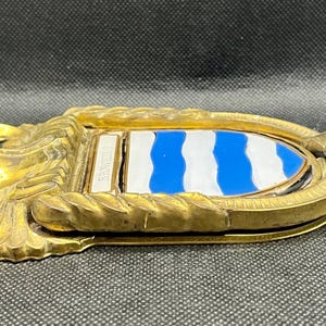 K&ouml;nnte beinhalten: Ein T&uuml;rklopfer aus Messing mit einem schildf&ouml;rmigen Emblem, das horizontale blaue und wei&szlig;e Wellenstreifen aufweist. Der Metallrahmen hat eine seilartige Textur und das Wort BROWNING ist auf einer kleinen rechteckigen Messingplatte eingraviert.