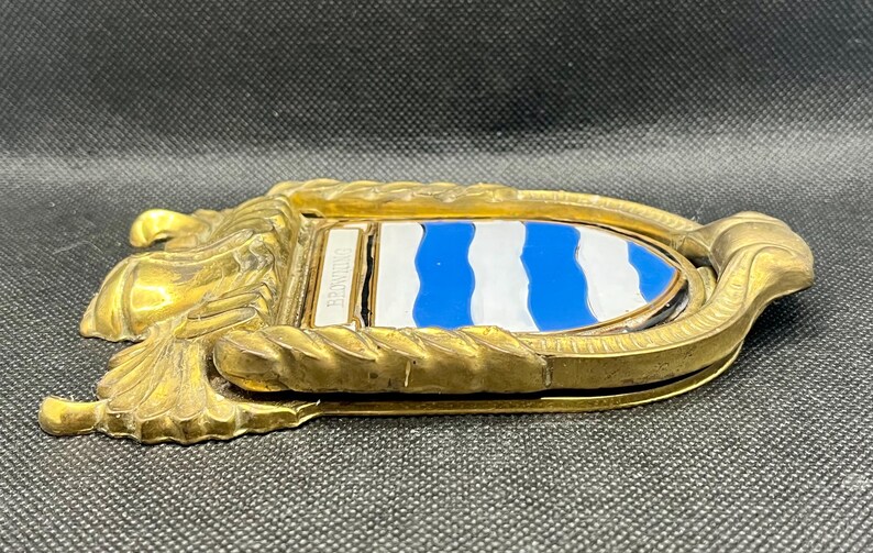 K&ouml;nnte beinhalten: Ein T&uuml;rklopfer aus Messing mit einem schildf&ouml;rmigen Emblem, das horizontale blaue und wei&szlig;e Wellenstreifen aufweist. Der Metallrahmen hat eine seilartige Textur und das Wort BROWNING ist auf einer kleinen rechteckigen Messingplatte eingraviert.