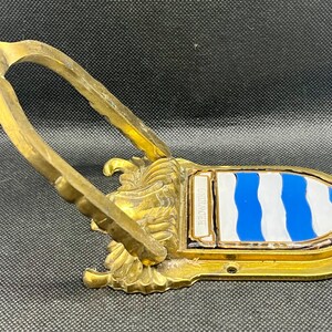 K&ouml;nnte beinhalten: Ein T&uuml;rklopfer aus Messing mit einem klappbaren U-f&ouml;rmigen Griff an einer dekorativen Basis. Die Basis zeigt ein Wappenschild mit abwechselnd blauen und wei&szlig;en Wellenstreifen sowie den Schriftzug BROWNING auf einer kleinen rechteckigen Platte.
