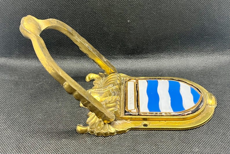 K&ouml;nnte beinhalten: Ein T&uuml;rklopfer aus Messing mit einem klappbaren U-f&ouml;rmigen Griff an einer dekorativen Basis. Die Basis zeigt ein Wappenschild mit abwechselnd blauen und wei&szlig;en Wellenstreifen sowie den Schriftzug BROWNING auf einer kleinen rechteckigen Platte.