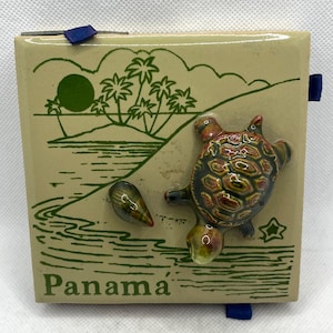 May include: A square ceramic tile featuring a green line drawing of a tropical beach scene with palm trees and a sun. A three-dimensional ceramic turtle and a small seashell are attached to the surface. The word Panama is printed in green text.