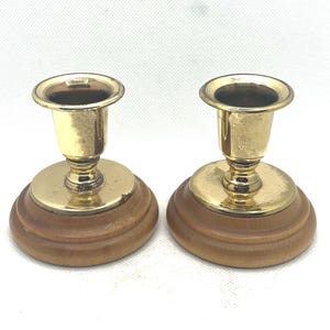May include: Two vintage candle holders. Each has a gold-colored metal cup and base, set atop a round, wooden base. The wood has a light brown color. The candle holders are designed to hold a single candle.
