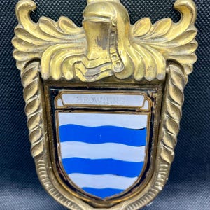 K&ouml;nnte beinhalten: Ein T&uuml;rklopfer aus Messing mit einem schildf&ouml;rmigen Emblem, das den Namen BROWNING in schwarzer Schrift tr&auml;gt. Der Schild zeigt horizontale blaue und wei&szlig;e Wellenstreifen. Der Metallrahmen ist mit kunstvollen Verzierungen und einem Helm verziert.