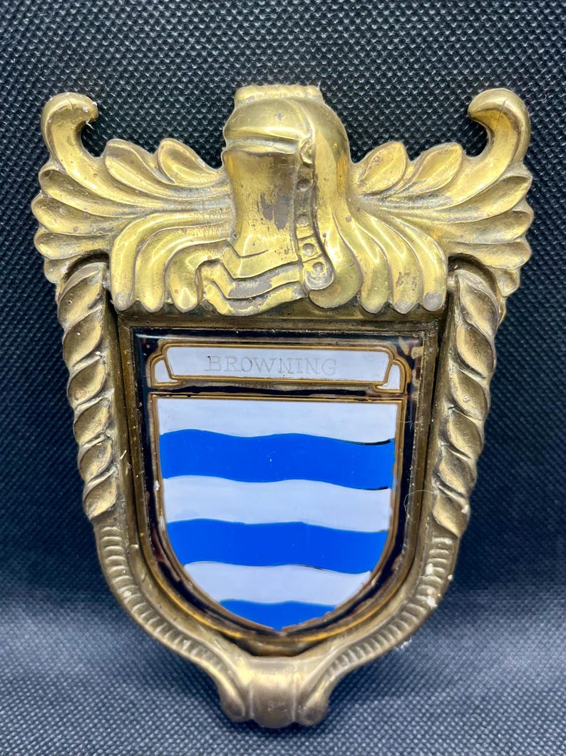 K&ouml;nnte beinhalten: Ein T&uuml;rklopfer aus Messing mit einem schildf&ouml;rmigen Emblem, das den Namen BROWNING in schwarzer Schrift tr&auml;gt. Der Schild zeigt horizontale blaue und wei&szlig;e Wellenstreifen. Der Metallrahmen ist mit kunstvollen Verzierungen und einem Helm verziert.