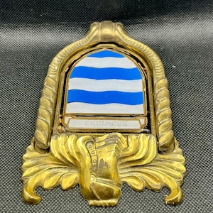 K&ouml;nnte beinhalten: Ein T&uuml;rklopfer aus Messing mit einem bogenf&ouml;rmigen Rahmen, einer seilartigen Umrandung und verziertem Schn&ouml;rkelwerk an der Basis. In der Mitte befindet sich ein Schild mit horizontalen blauen und wei&szlig;en Wellenstreifen und einem Banner mit BROWNING.