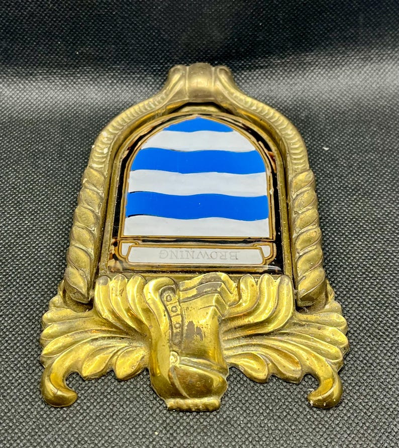 K&ouml;nnte beinhalten: Ein T&uuml;rklopfer aus Messing mit einem bogenf&ouml;rmigen Rahmen, einer seilartigen Umrandung und verziertem Schn&ouml;rkelwerk an der Basis. In der Mitte befindet sich ein Schild mit horizontalen blauen und wei&szlig;en Wellenstreifen und einem Banner mit BROWNING.