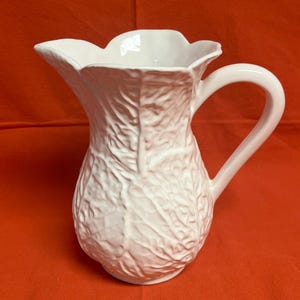 May include: A white ceramic pitcher featuring a textured surface that mimics the appearance of overlapping cabbage leaves. The vessel has a wide, scalloped rim, a curved handle on the right side, and a prominent spout for pouring liquids.