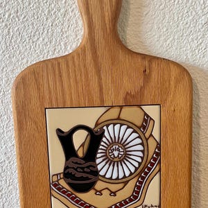 May include: Wooden cutting board with a decorative tile insert. The tile features a black vase, a white and brown wheel design, and a tan background with red and brown accents. The board has a handle with a hole for hanging.