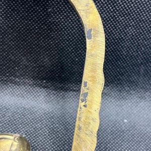 K&ouml;nnte beinhalten: Ein gebogener Messinggriff mit einer texturierten, gewellten Au&szlig;enkante und einer glatten Innenfl&auml;che. Das Metall weist Gebrauchsspuren mit kleinen dunklen Oxidationsflecken und sichtbaren Kratzern entlang des goldfarbenen Metallrahmens auf.