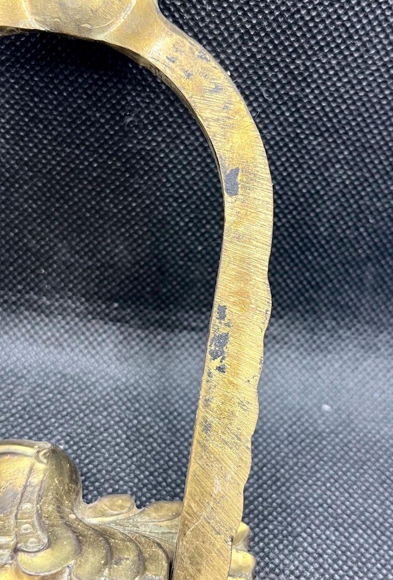 K&ouml;nnte beinhalten: Ein gebogener Messinggriff mit einer texturierten, gewellten Au&szlig;enkante und einer glatten Innenfl&auml;che. Das Metall weist Gebrauchsspuren mit kleinen dunklen Oxidationsflecken und sichtbaren Kratzern entlang des goldfarbenen Metallrahmens auf.