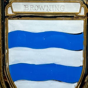 K&ouml;nnte beinhalten: Eine rechteckige Metallplakette mit einem wei&szlig;en Emaille-Schild, das drei horizontale blaue Wellenlinien aufweist. &Uuml;ber dem Schild befindet sich ein wei&szlig;es Banner mit dem Wort BROWNING in Gro&szlig;buchstaben. Das St&uuml;ck ist in einen goldfarbenen Metallrahmen gefasst.