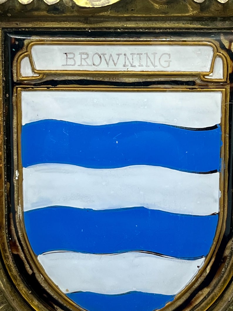 K&ouml;nnte beinhalten: Eine rechteckige Metallplakette mit einem wei&szlig;en Emaille-Schild, das drei horizontale blaue Wellenlinien aufweist. &Uuml;ber dem Schild befindet sich ein wei&szlig;es Banner mit dem Wort BROWNING in Gro&szlig;buchstaben. Das St&uuml;ck ist in einen goldfarbenen Metallrahmen gefasst.