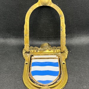 K&ouml;nnte beinhalten: Ein T&uuml;rklopfer aus Messing mit einem bogenf&ouml;rmigen Griff und einem zentralen schildf&ouml;rmigen Emblem. Der Schild zeigt horizontale blaue und wei&szlig;e Streifen mit dem Namen BROWNING in wei&szlig;er Schrift auf einem kleinen horizontalen Balken.