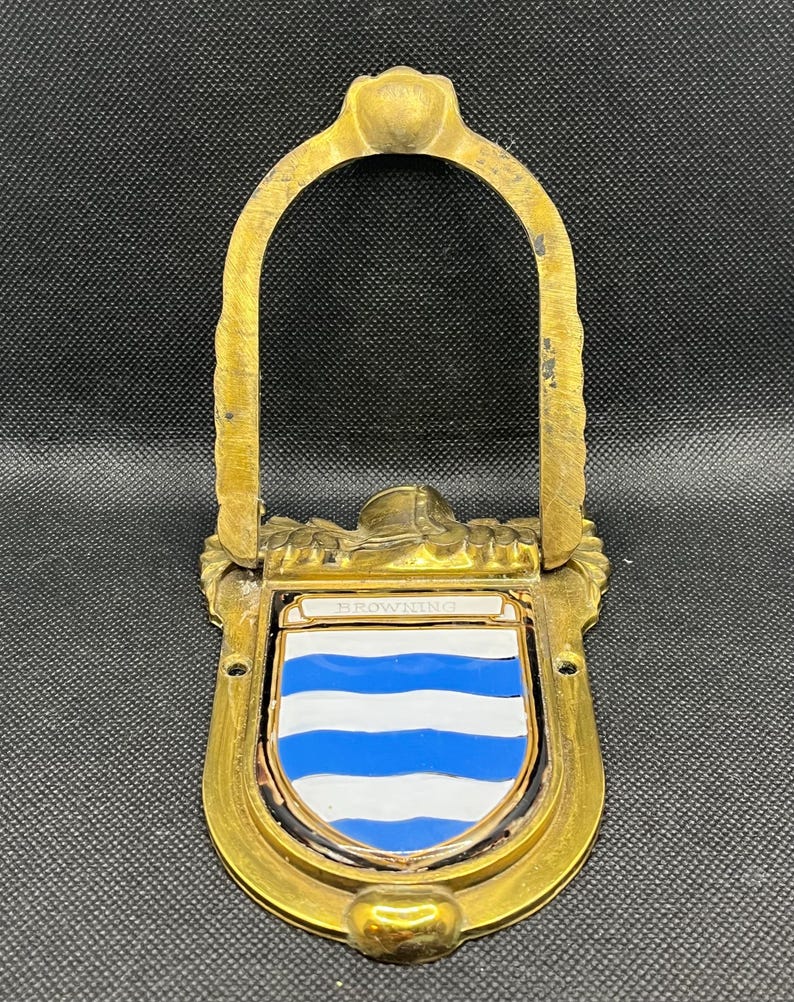 K&ouml;nnte beinhalten: Ein T&uuml;rklopfer aus Messing mit einem bogenf&ouml;rmigen Griff und einem zentralen schildf&ouml;rmigen Emblem. Der Schild zeigt horizontale blaue und wei&szlig;e Streifen mit dem Namen BROWNING in wei&szlig;er Schrift auf einem kleinen horizontalen Balken.