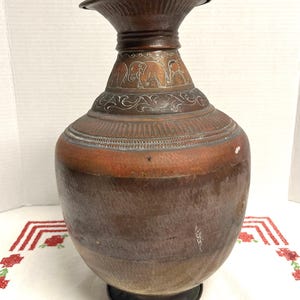 May include: A copper-toned metal vase with a wide, flared rim and a rounded body. The neck features an engraved band depicting a series of elephants, complemented by intricate scrollwork and linear patterns etched into the metal surface.