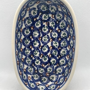 May include: An oval ceramic bowl featuring a repeating pattern of blue sponge-painted circles, small green leaf-like motifs, and tiny brown accents on a cream-colored background. The interior of the bowl is fully decorated with this pattern.