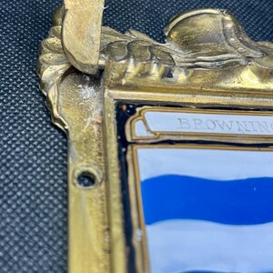 K&ouml;nnte beinhalten: Eine messingfarbene Metallplakette mit verziertem Oberrand und einem zentralen Schildmotiv. Das Schild zeigt horizontale blaue und wei&szlig;e Streifen mit dem Text BROWNING oben. Der Metallrahmen weist eine verwitterte Oberfl&auml;che auf.