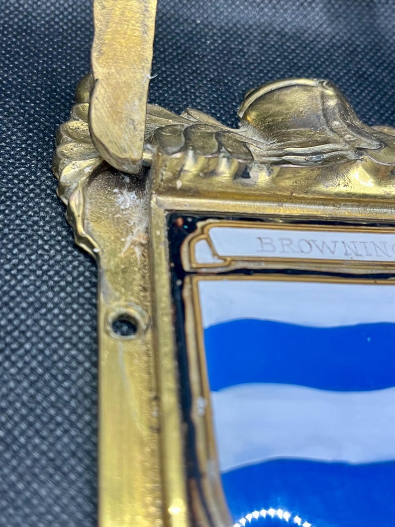 K&ouml;nnte beinhalten: Eine messingfarbene Metallplakette mit verziertem Oberrand und einem zentralen Schildmotiv. Das Schild zeigt horizontale blaue und wei&szlig;e Streifen mit dem Text BROWNING oben. Der Metallrahmen weist eine verwitterte Oberfl&auml;che auf.