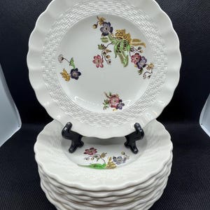 May include: A stack of white ceramic plates with a scalloped edge and a woven texture around the rim. Each plate features a floral design with purple, pink, and yellow accents. The plates are stacked on a black surface.