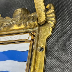 K&ouml;nnte beinhalten: Eine messingfarbene Metallplakette mit einem verzierten, skulptierten Rand und einem zentralen emaillierten Schild. Das Schild zeigt horizontale blaue und wei&szlig;e Streifen mit dem Text BROWNING auf einem wei&szlig;en Banner oben. Ein Montageloch ist sichtbar.