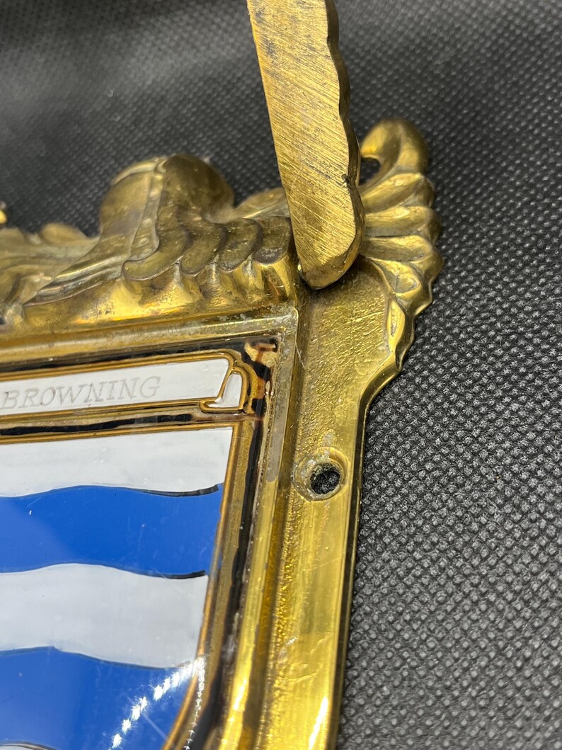 K&ouml;nnte beinhalten: Eine messingfarbene Metallplakette mit einem verzierten, skulptierten Rand und einem zentralen emaillierten Schild. Das Schild zeigt horizontale blaue und wei&szlig;e Streifen mit dem Text BROWNING auf einem wei&szlig;en Banner oben. Ein Montageloch ist sichtbar.