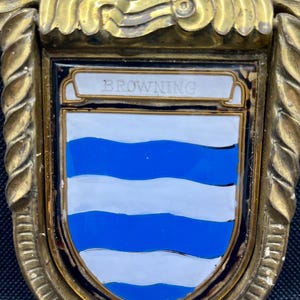 K&ouml;nnte beinhalten: Ein heraldischer Schild aus Messing mit einem zentralen Emaille-Wappen, das abwechselnd blaue und wei&szlig;e, gewellte horizontale Streifen aufweist. Das Wort BROWNING steht auf einem wei&szlig;en Banner oben auf dem Schild, der von einem gedrehten Metallrand umrahmt ist.
